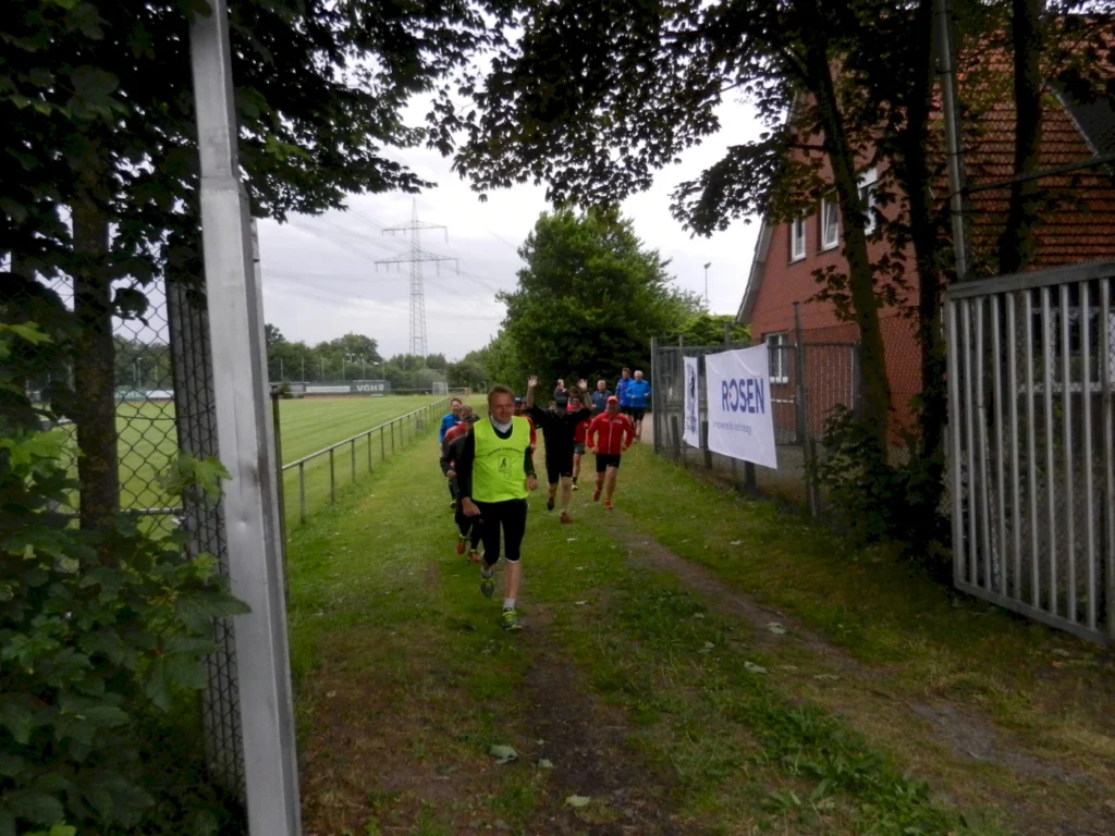 Läufer auf einem Trainingsweg neben einem Fußballfeld, umgeben von Bäumen und einer Zaunanlage, während sie an einem Sportevent teilnehmen.