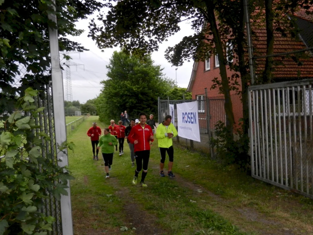 Gruppe von Menschen joggt auf einem Pfad neben einem Gebäude mit einem Werbebanner, von Bäumen gesäumt.