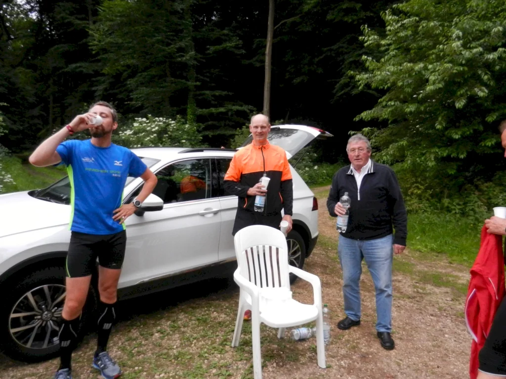 Drei Männer machen Trinkpause beim Laufen im Wald neben einem geparkten Auto, umgeben von Bäumen und Natur.