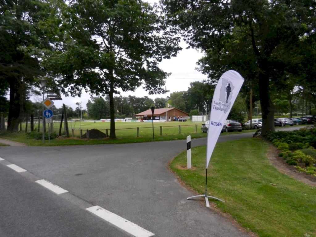 Eingang zu einem Sportgelände mit einem Vereinsgebäude im Hintergrund, umgeben von Bäumen und Parkplätzen. Ein Fahnenmast mit dem Logo der Laufgemeinschaft Emsland steht an der Straßeneinfahrt.