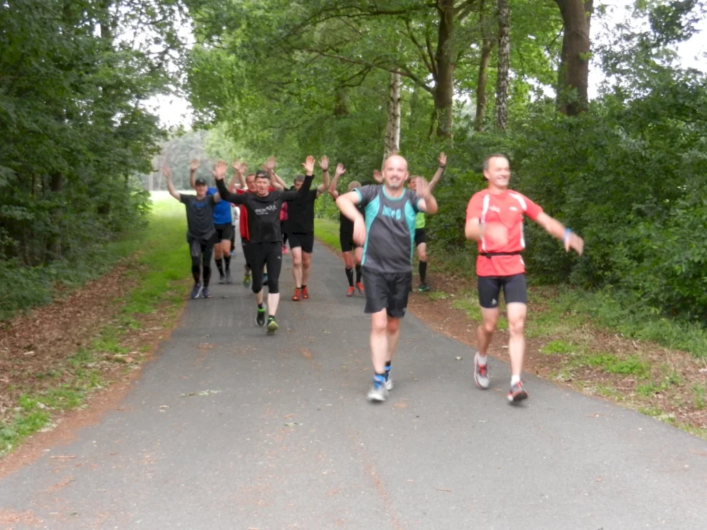 Gruppe von Läufern auf einer Waldstraße, freut sich mit erhobenen Händen beim gemeinsamen Lauf im Freien.