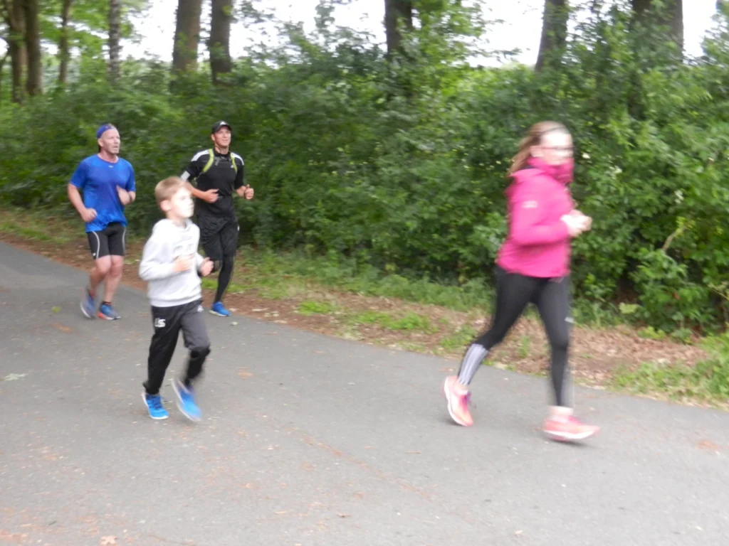 Gruppe von Menschen joggt auf einem Waldweg, darunter ein Kind, in sportlicher Kleidung und unterschiedlichem Tempo.