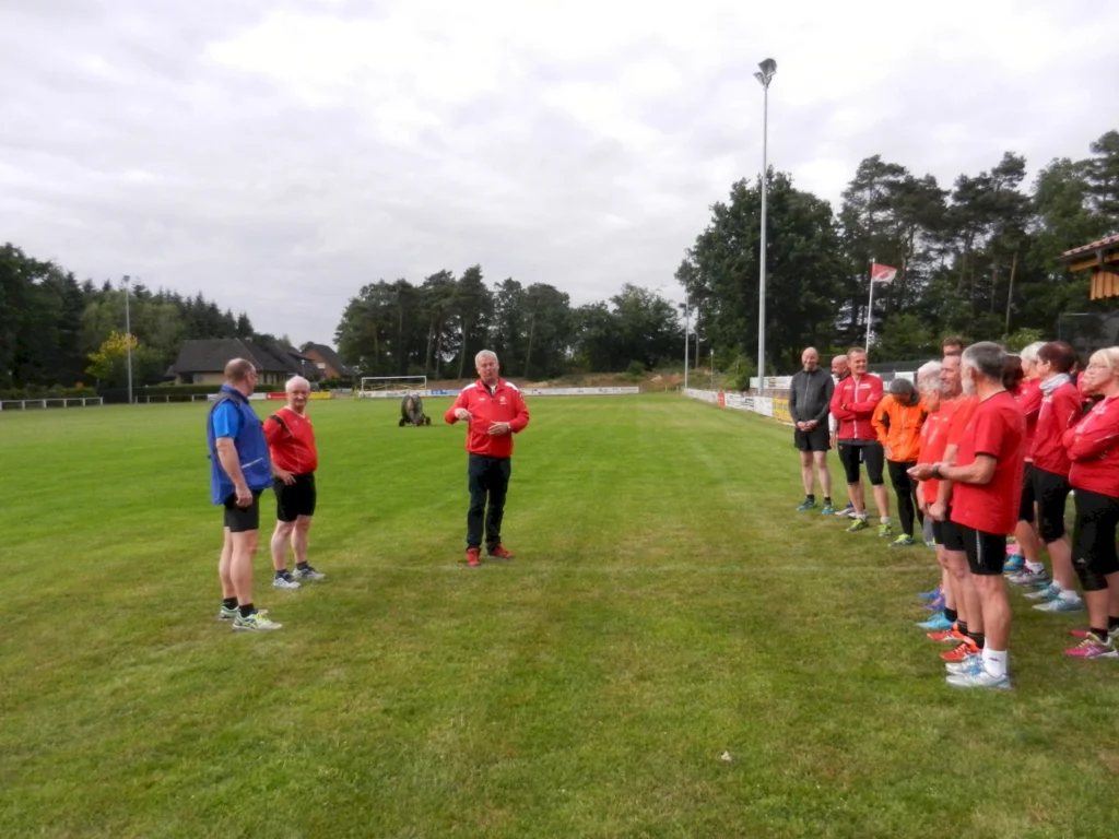Gruppe von Menschen in Sportkleidung auf einem grünen Fußballplatz bei einer Besprechung im Freien, bewölkter Himmel.