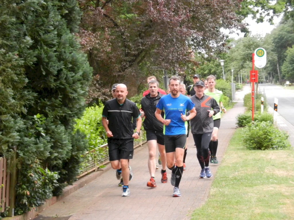 Gruppe von Männern joggt auf einem Gehweg neben einer Straße, umgeben von Bäumen und grüner Landschaft.