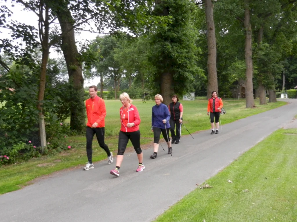 Gruppe von fünf Menschen in Sportkleidung, die auf einem Weg in einem Park spazieren gehen, umgeben von Bäumen und Grünflächen. Ideal für Fitness und Outdoor-Aktivitäten.