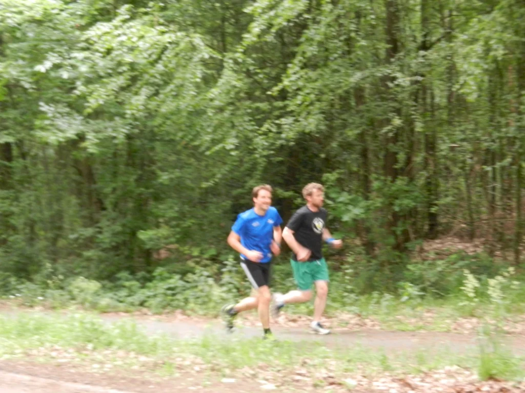 Zwei Männer joggen auf einem Waldweg, umgeben von grünen Bäumen und Pflanzen. Der Mann links trägt ein blaues Shirt und kurze Hosen, der Mann rechts ein schwarzes Shirt und grüne Shorts.