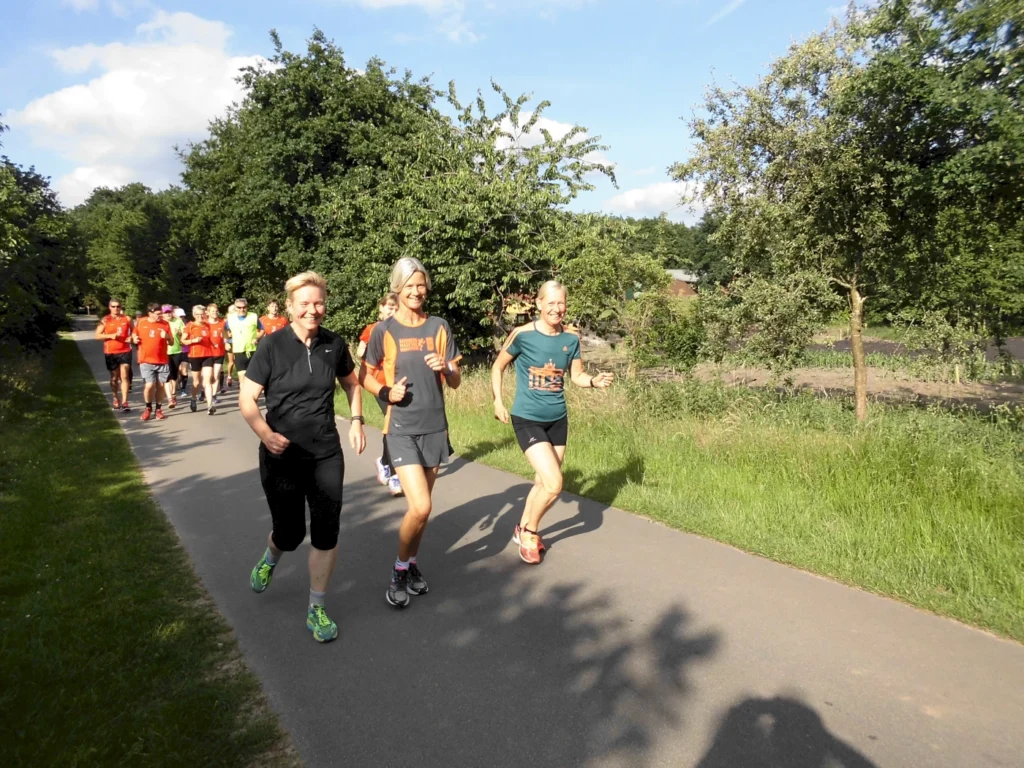 Läufergruppe auf einem Waldweg, bestehend aus mehreren Teilnehmern in sportlicher Kleidung, die gemeinsam joggen. Zwei Frauen führen die Gruppe an, lächeln und zeigen Daumen hoch. Umgebende Natur mit Bäumen und grünem Gras. Ideal für Fitness- und Laufveranstaltungen.