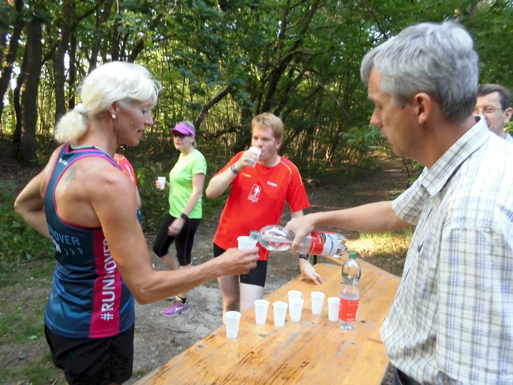 Läuferin erhält Wasser von einem Helfer während einer Laufveranstaltung im Wald. Im Hintergrund stehen weitere Teilnehmer in sportlicher Kleidung, die ebenfalls Wasser trinken. Die Szene zeigt die Bedeutung der Hydration beim Sport.