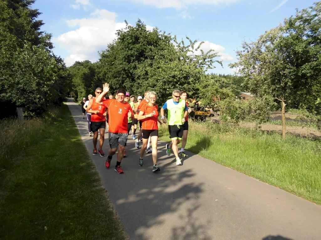 Gruppe von Läufern in roten Trikots joggt auf einem sonnigen Weg durch grüne Landschaft, ein Läufer winkt.