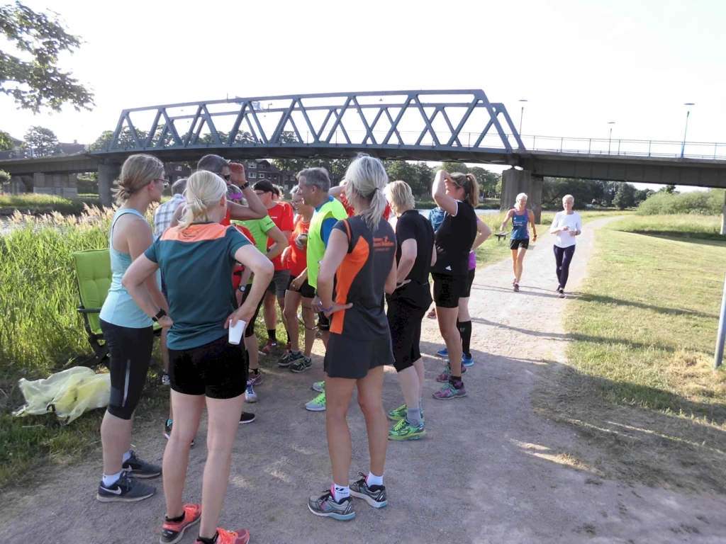 Gruppe von Menschen in Sportbekleidung versammelt sich auf einem Weg neben einer Brücke an einem sonnigen Tag.