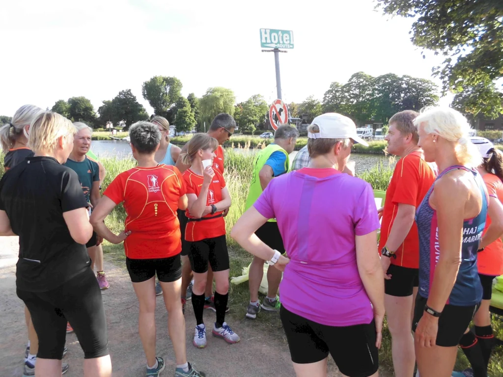 Gruppe von Läufern in Sportbekleidung, die sich am Ufer eines Gewässers unterhalten, während ein Hotelzeichen im Hintergrund sichtbar ist. Ideal für Themen wie Laufen, Fitness und Outdoor-Aktivitäten.