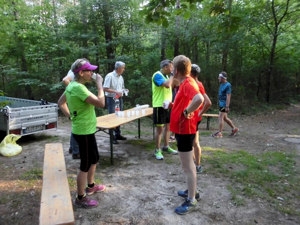 Läufergruppe bei Erfrischungspause im Wald, trinken Wasser an einem Tisch, umgeben von Bäumen.