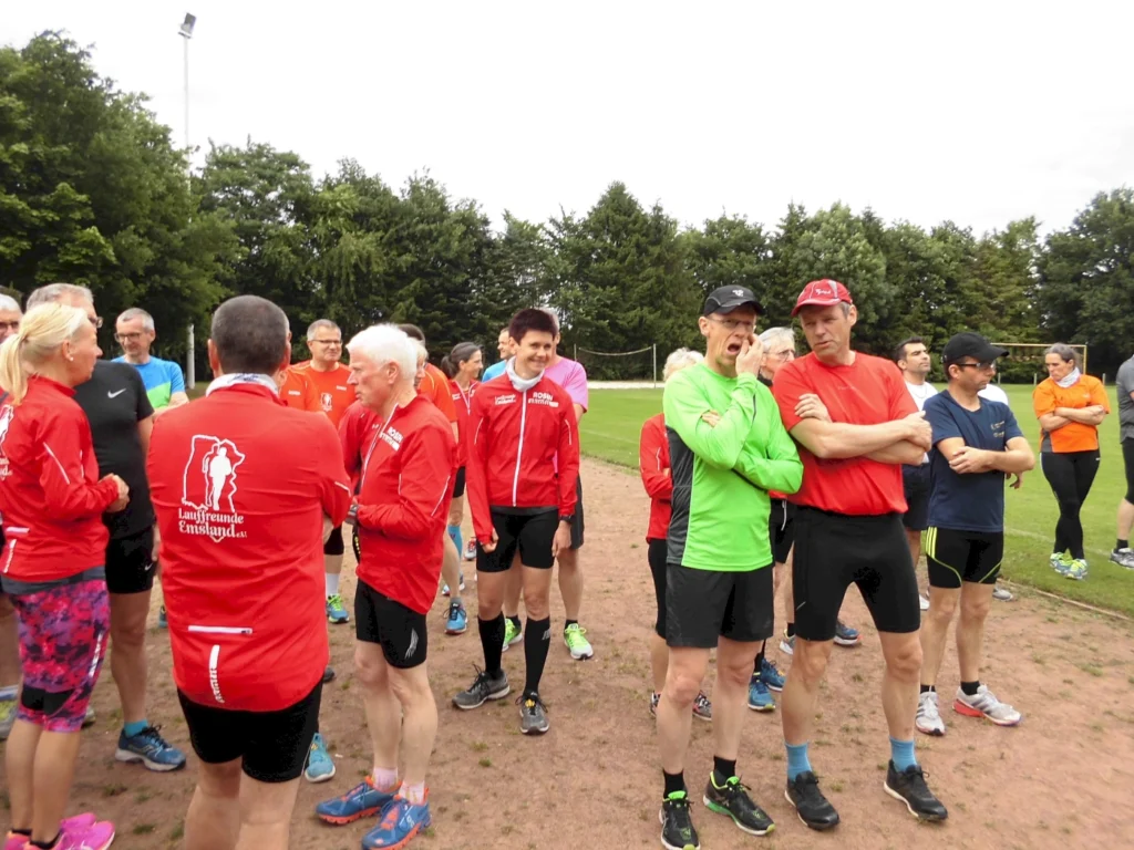 Gruppentreffen der Laufgruppe „Lauf Freunde Emstal“ auf einem Sportplatz, Teilnehmer in Laufkleidung, darunter rote und grüne Shirts, stehen zusammen und unterhalten sich, umgeben von Bäumen und Sporteinrichtungen.