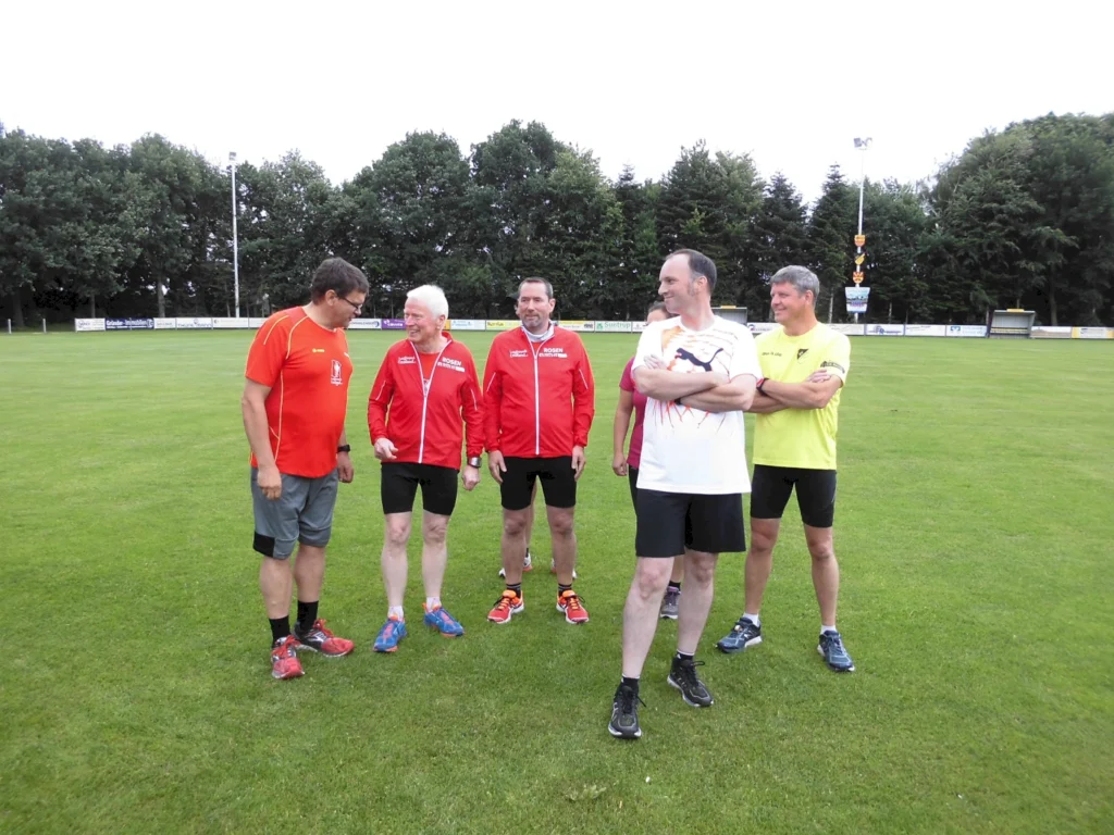 Fünf Männer in Sportkleidung stehen auf einem Fußballfeld und unterhalten sich. Im Hintergrund sind Bäume und eine Sportanlage sichtbar.