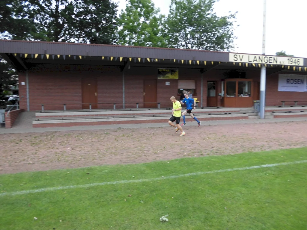 Zwei junge Fußballspieler laufen auf dem Sportplatz des SV Langen 1946, während im Hintergrund die Tribüne mit Sitzplätzen und Türen zu sehen ist. Grüne Rasenfläche und Bäume umgeben das Gelände.