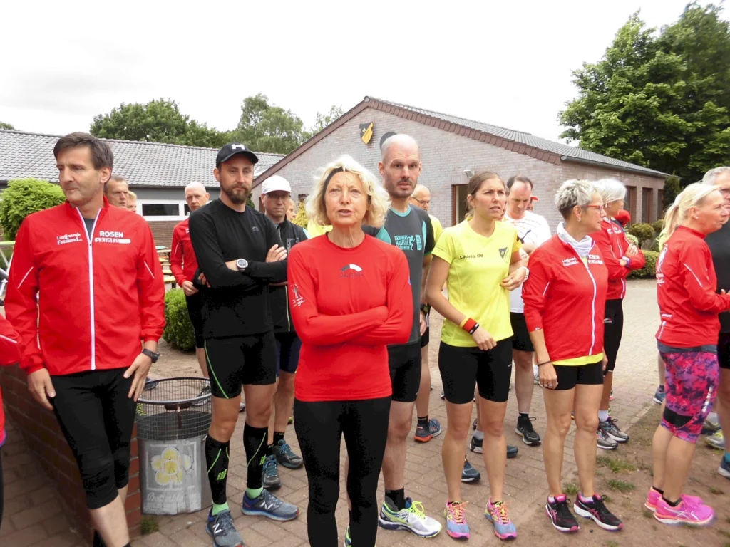 Gruppe von Läufern in Sportkleidung, die sich vor einem Gebäude versammelt haben, bereit für ein Rennen oder eine Laufveranstaltung. Einige tragen rote Jacken, andere schwarze und gelbe Shirts. Die Szene zeigt eine Mischung aus Männern und Frauen, die auf den Start warten.