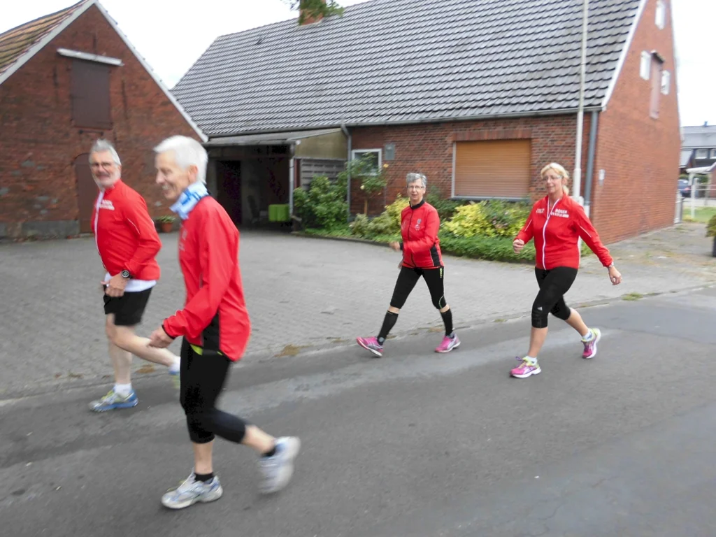 Vier Personen in roten Laufjacken joggen auf einer Straße in einem Wohngebiet. Im Hintergrund sind Häuser und grüne Pflanzen sichtbar. Die Gruppe zeigt eine aktive Lebensweise und Freude am Sport.