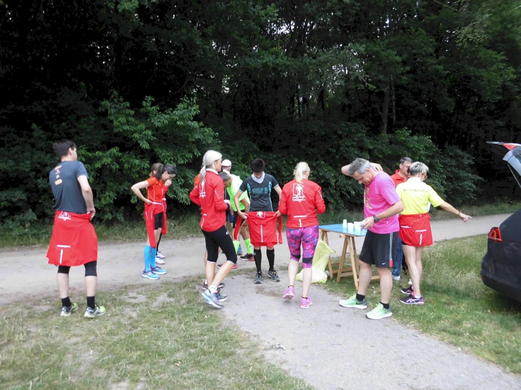 Gruppe von Läufern in sportlicher Kleidung bei einer Laufveranstaltung, die sich um einen Tisch mit Getränken versammelt. Im Hintergrund Bäume und ein unbefestigter Weg.