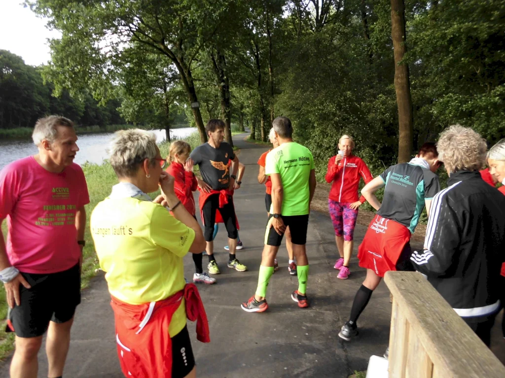 Gruppe von Läufern in bunten Sportoutfits, die sich am Ufer eines Gewässers aufwärmen oder besprechen, umgeben von Bäumen und Natur. Ideal für Themen wie Laufsport, Fitness und Outdoor-Aktivitäten.