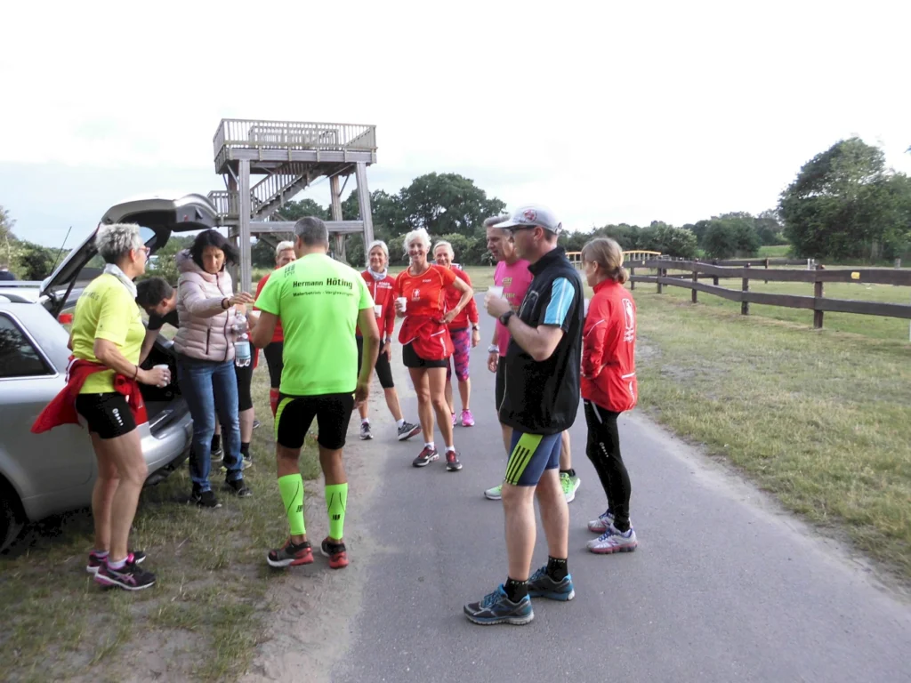Gruppe von Läufern in Sportkleidung, die sich vor einem Auto versammeln, während eine Frau etwas aus dem Kofferraum holt. Im Hintergrund ist ein Aussichtsturm und eine grüne Landschaft zu sehen.