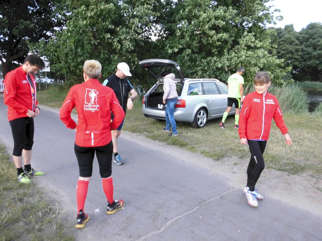 Gruppentreffen von Laufbegeisterten in roten Jacken an einem ruhigen Weg, während sie sich auf einen Lauf vorbereiten. Im Hintergrund ist ein silberner Pkw zu sehen und die Umgebung ist von Bäumen und Gras umgeben.