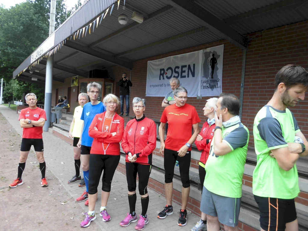 Laufgruppe vor einer Tribüne mit Sportkleidung, im Gespräch und bereit für ein Event. Hintergrund: Banner und Bäume.