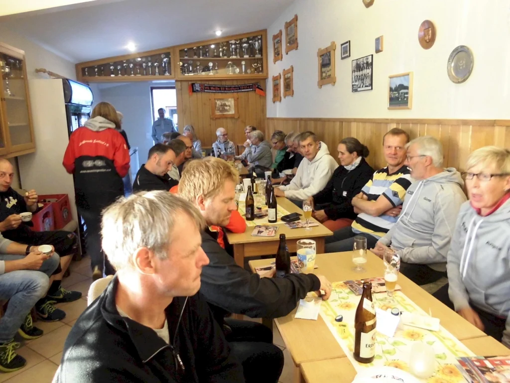 Gesellige Runde in einem Gasthaus: Männer und Frauen sitzen an Tischen, trinken Bier und genießen Snacks, während sie sich unterhalten. Die Wände sind mit Bildern und Trophäen dekoriert, was eine einladende Atmosphäre schafft.