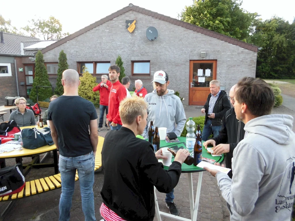 Gesellige Runde von Menschen bei einer Veranstaltung im Freien, umgeben von Tischen und Getränken, vor einem Gebäude mit einem Schild. Die Teilnehmer, darunter Männer und Frauen, unterhalten sich und genießen die Atmosphäre.
