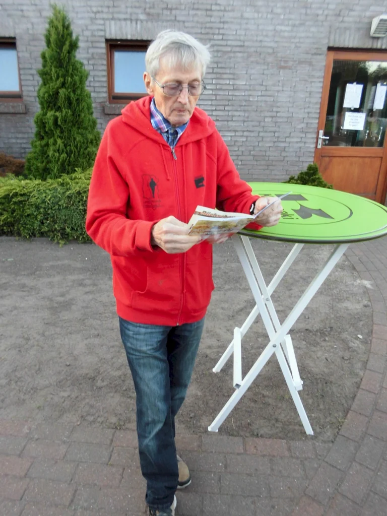 Älterer Mann in rotem Hoodie steht an einem Tisch und liest eine Broschüre im Freien, umgeben von Grünpflanzen und einer Backsteinmauer.