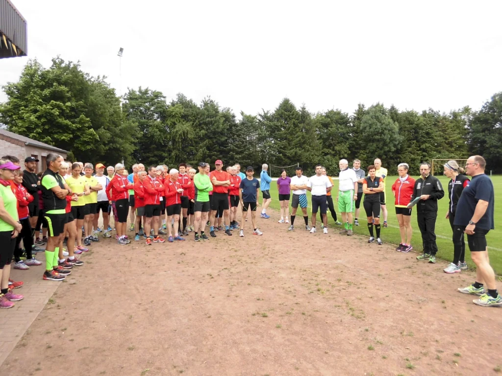 Gruppenfoto von Teilnehmern eines Sportevents im Freien, die in sportlicher Kleidung versammelt sind. Die Gruppe besteht aus Männern und Frauen unterschiedlichen Alters, die auf einem Sportplatz stehen und einem Redner zuhören. Im Hintergrund sind Bäume und ein Sportfeld sichtbar.