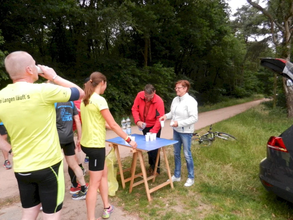 Läufer in gelben T-Shirts nehmen an einem Wettkampf teil, während sie an einem Verpflegungstisch mit Wasserflaschen und Bechern vorbeigehen. Im Hintergrund sind Bäume und ein Fahrrad zu sehen.
