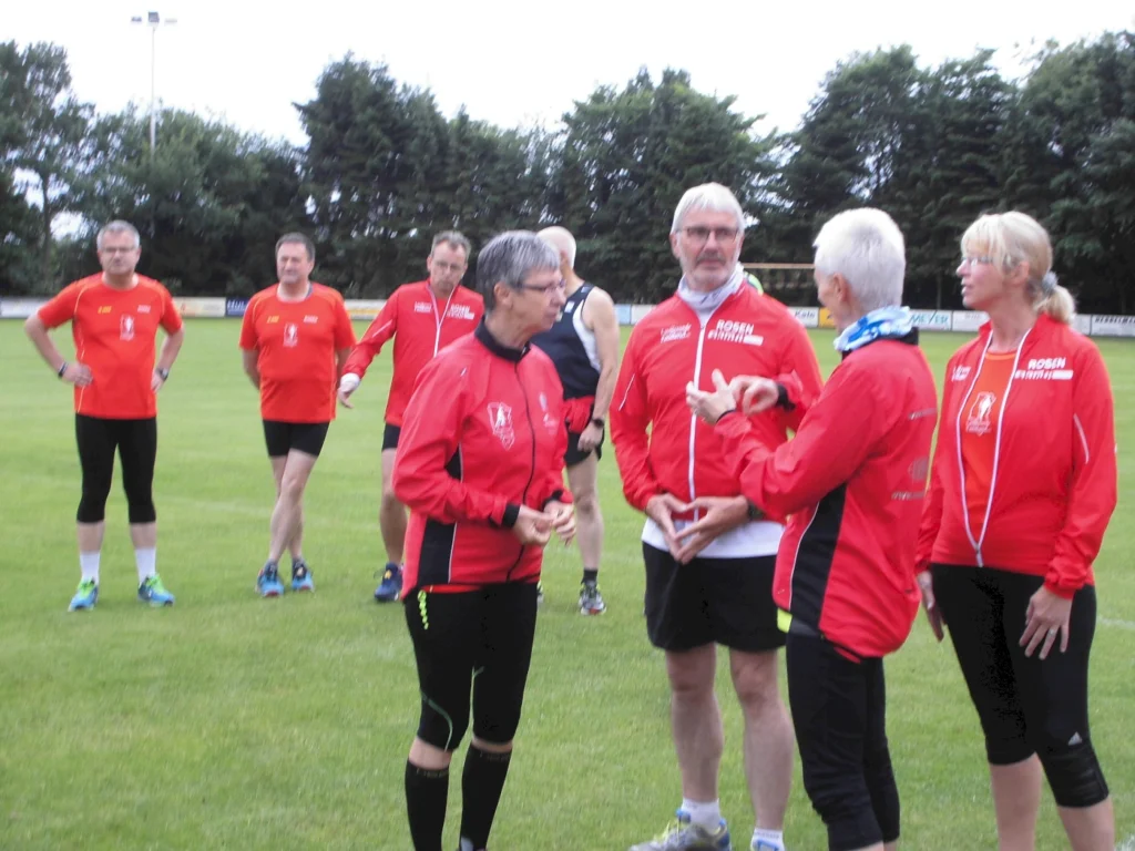 Gruppe von Läufern in roten Trainingsjacken diskutiert auf einem Sportfeld im Freien.