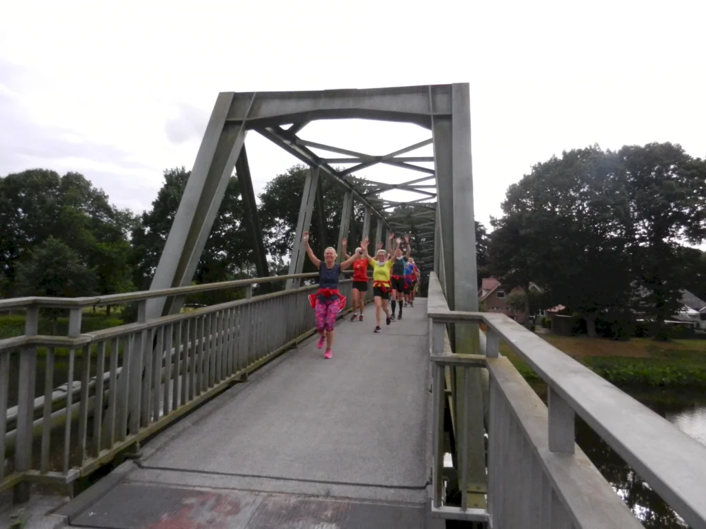 Läufergruppe auf einer Brücke, die fröhlich die Arme hebt, umgeben von grüner Landschaft und Häusern im Hintergrund. Ideal für Sport- und Freizeitaktivitäten.