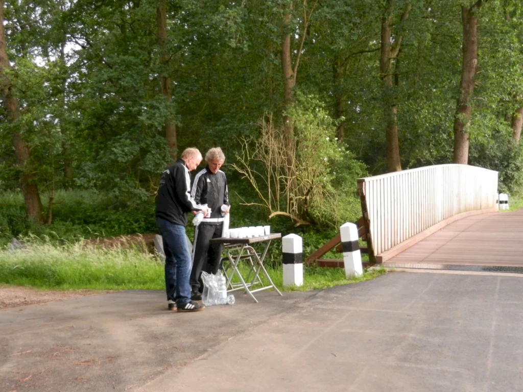 Zwei Männer bereiten an einem Tisch mit Getränken vor einer Holzbrücke in einem Waldgebiet vor. Die Umgebung ist grün und sonnig, mit Bäumen im Hintergrund.