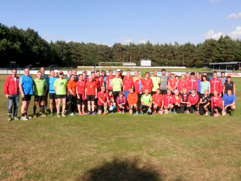 Eine Gruppe von Menschen in bunter Sportkleidung posiert auf einem Sportplatz. Im Hintergrund ist Wald zu sehen.