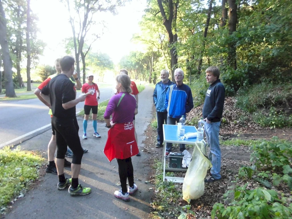 Läufergruppe und Helfer an einem Versorgungspunkt im Wald entlang einer Straße, sonniges Wetter, grüne Umgebung.