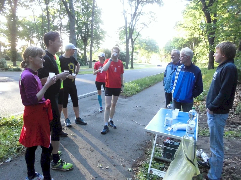 Gruppe von Läufern und Unterstützern an einer Verpflegungsstation während eines Lauf-Events in der Natur, umgeben von Bäumen und einer Straße. Die Teilnehmer trinken Wasser und unterhalten sich.
