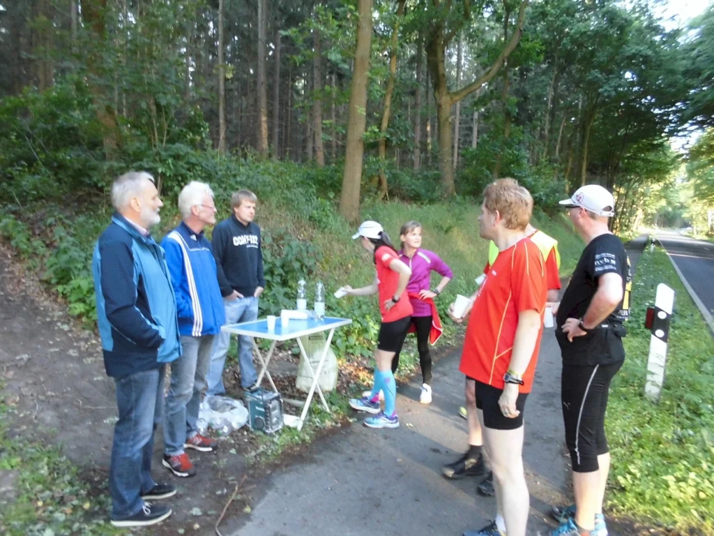 Läufer und Helfer an einem Verpflegungspunkt im Wald während eines Laufevents, umgeben von Bäumen und einer Straße.