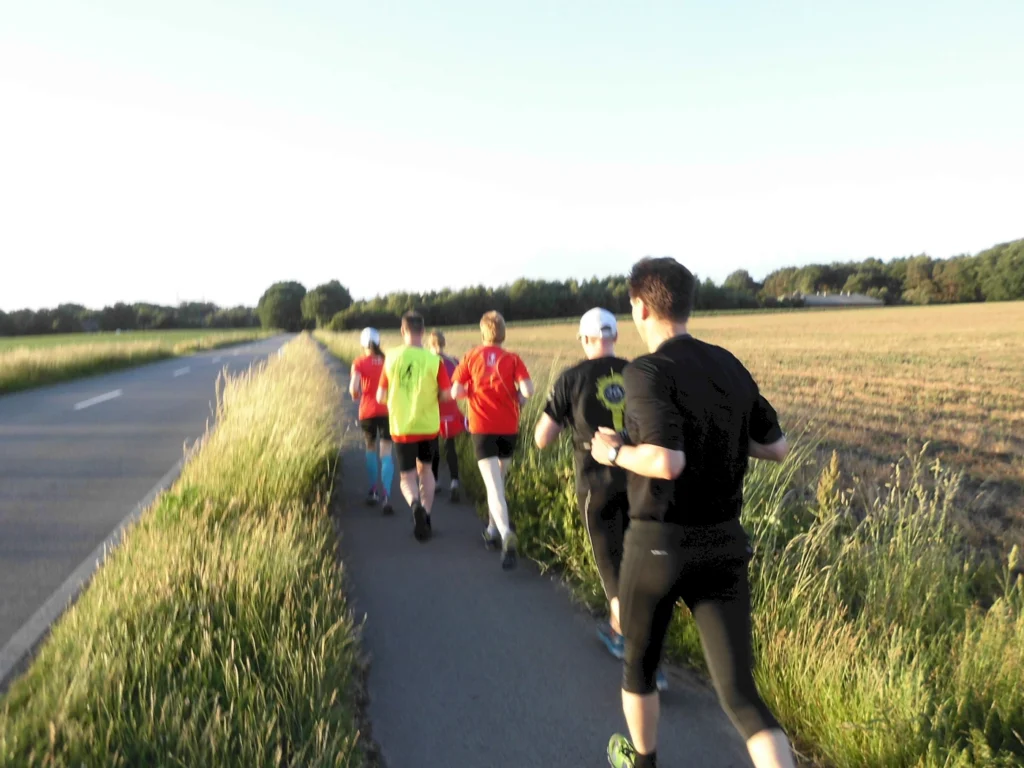 Gruppe von Menschen joggt auf einem ländlichen Weg bei Sonnenuntergang, neben einem Feld und einer Straße.