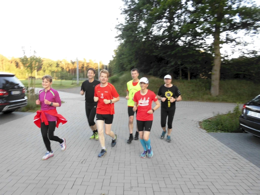 Gruppe von sechs Personen joggt in sportlicher Kleidung auf einer mit Bäumen gesäumten Straße bei Tageslicht.