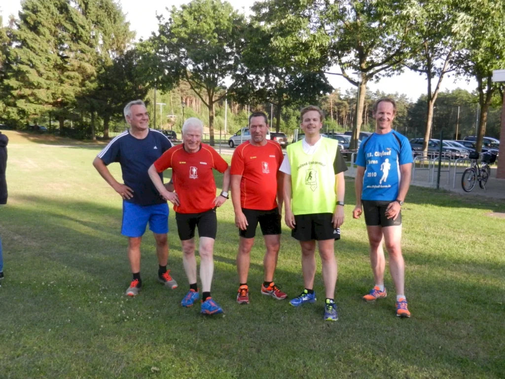 Fünf Männer in Sportbekleidung stehen auf einer Wiese, lächeln und posieren nach dem Laufen im Park.