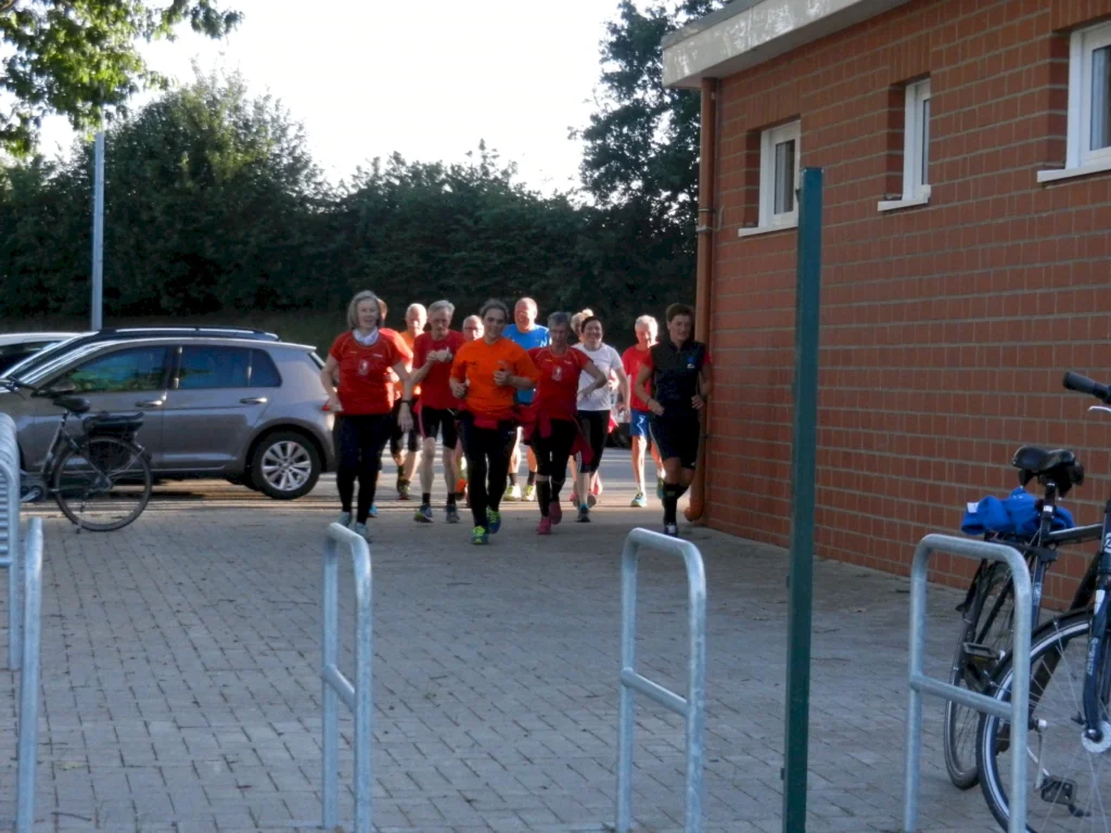 Gruppe von Läufern in roten und orangefarbenen Shirts, die in einem Außenbereich joggen, während ein Auto und Fahrräder im Hintergrund sichtbar sind.
