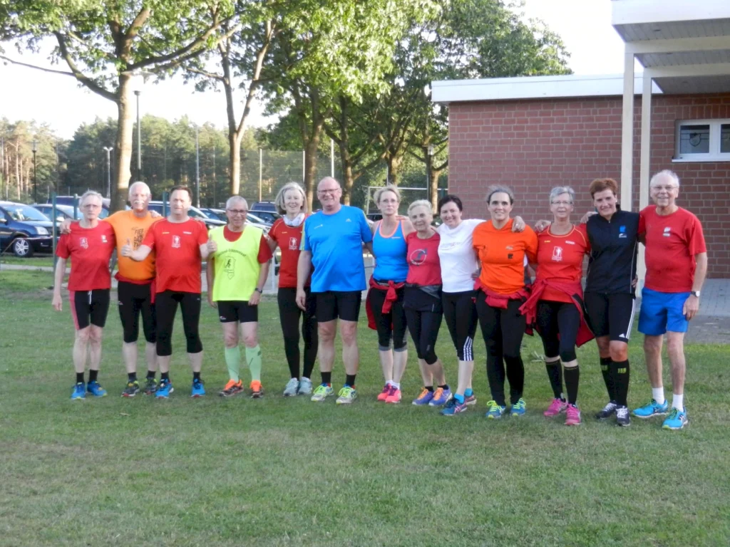 Gruppe von Läufern in Sportkleidung posiert auf einer Wiese vor einem Gebäude und Bäumen an einem sonnigen Tag.