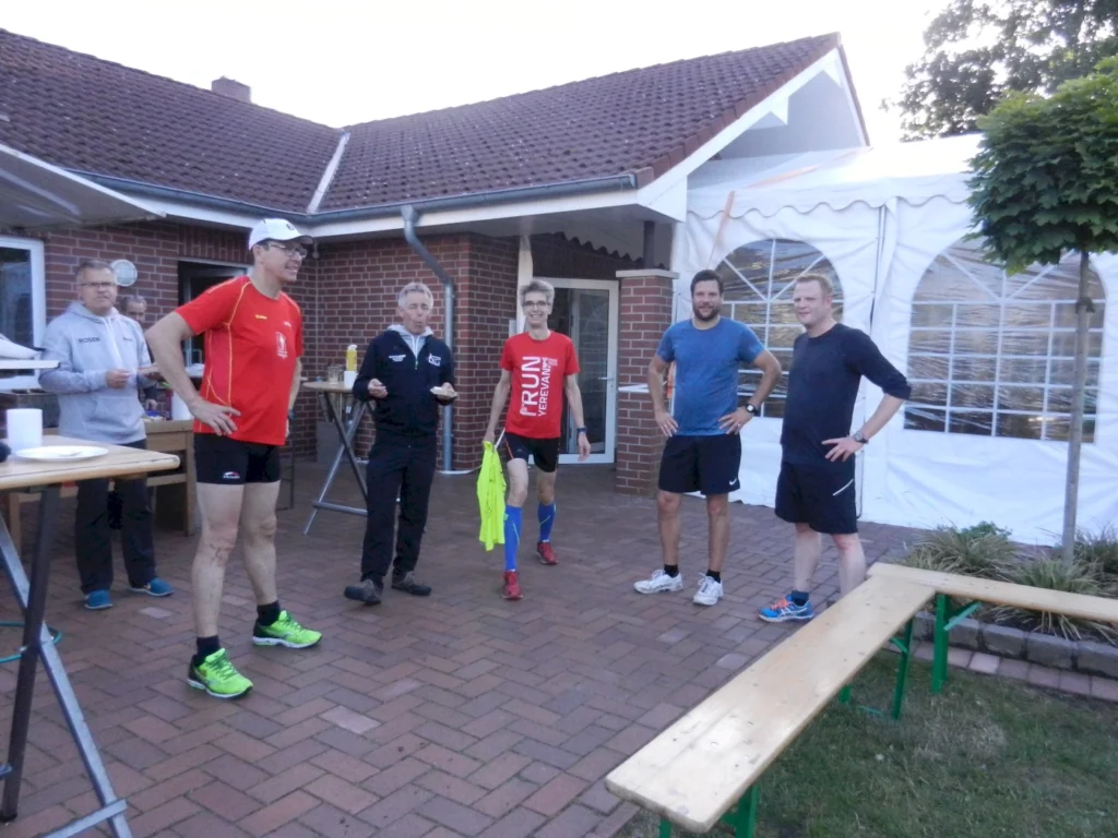 Gruppe von Männern in Sportkleidung steht auf einer Terrasse, bereit für ein Lauf-Event bei einem Haus mit Zelt im Hintergrund.