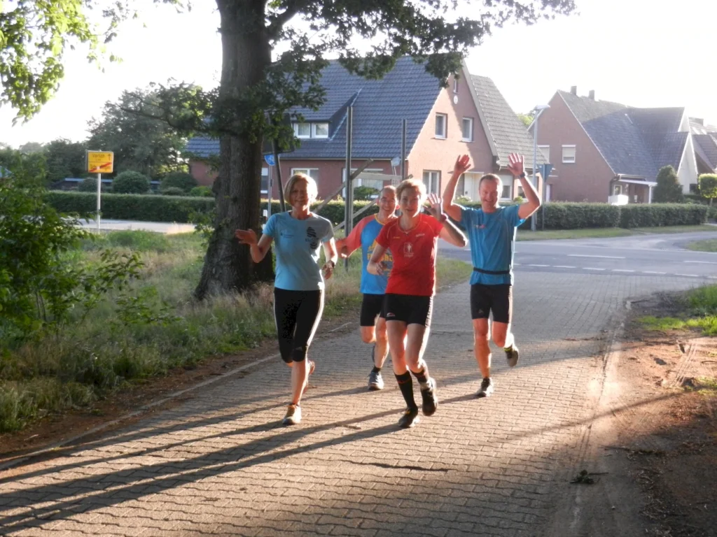 Vier Läufer joggen auf einem ländlichen Weg im Sonnenlicht, lächelnd und mit erhobenen Armen.