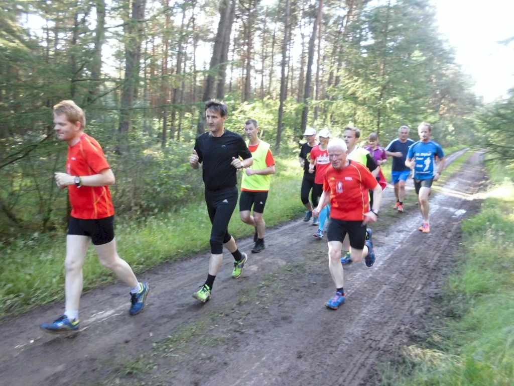 Gruppe von Läufern joggt im Wald auf einem schlammigen Weg, umgeben von Bäumen bei Tageslicht.