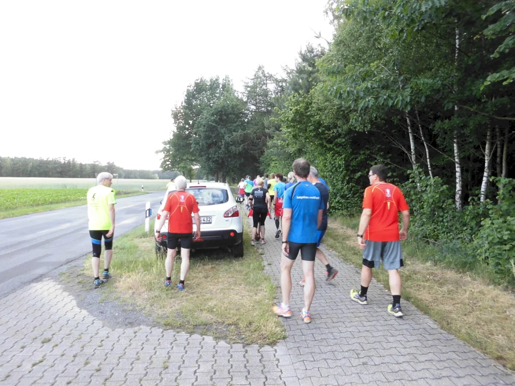Gruppe von Joggern in bunten Sportoutfits läuft auf einem Waldweg. Auto parkt am Straßenrand. Sommerliche Szene.