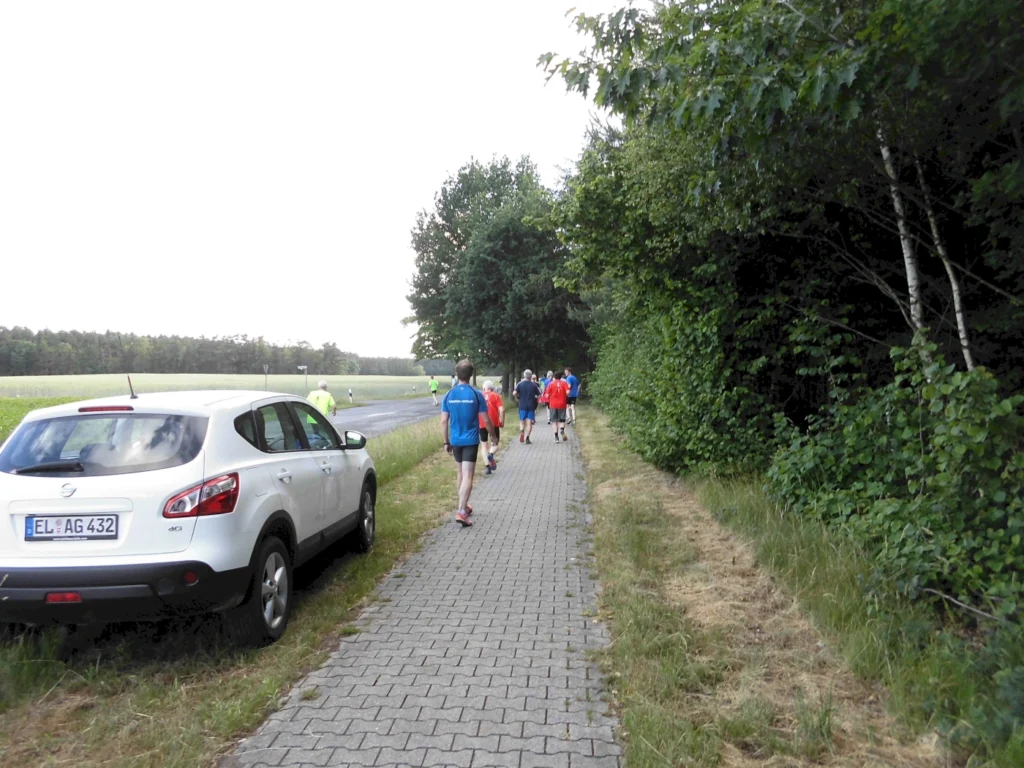 Gruppe von Menschen joggt auf Waldweg neben der Straße, während ein weißes Auto am Rand geparkt ist.