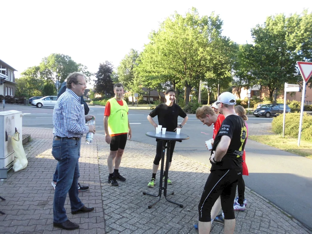 Gruppe von Menschen steht um einen Stehtisch im Freien, trägt Sportkleidung, im Hintergrund eine Straße und Bäume.