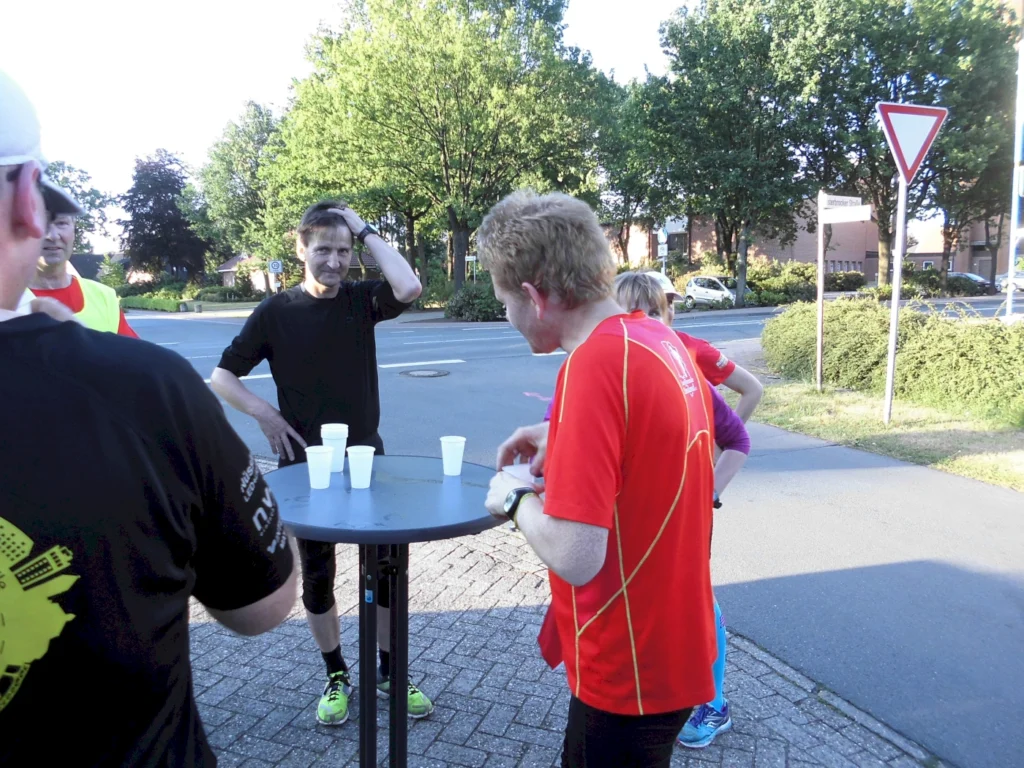 Gruppe von Läufern in sportlicher Kleidung, die an einem Tisch mit Bechern stehen, während sie sich in einer urbanen Umgebung in der Morgensonne unterhalten.